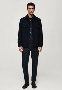 Donkerblauwe jas met twee borstzakken, gecombineerd met een bijpassende overhemd met knopen en slim-fit broek. Zwarte enkellaarzen maken de look compleet.