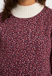 T-shirt à manches longues avec un col blanc côtelé. Tissu bordeaux avec un motif léopard en rose, crème et marine. Texture douce.