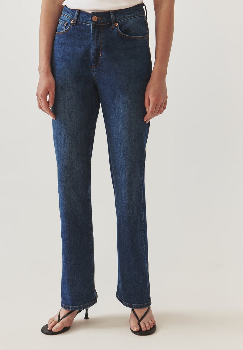 Jeans de mezclilla azul oscuro con cintura alta, corte recto y detalles de costura sutiles. Combinados con sandalias de tacón negras.