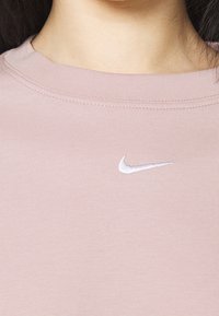 Ljusrosa bomullströja med rund hals och broderad vit Nike-logga på bröstet. Tyget har en mjuk, slät yta.
