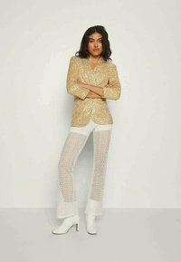Woman standing with arms crossed wearing a gold sequin blazer, white textured pants, and white heeled boots against plain background.