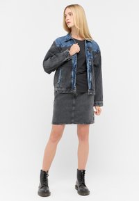 Schwarze Jeansjacke mit blauen Akzenten, kombiniert mit einem eng anliegenden schwarzen Oberteil und einem grauen Jeansrock. Das Model trägt schwarze Schnürstiefel im Kampfstil.
