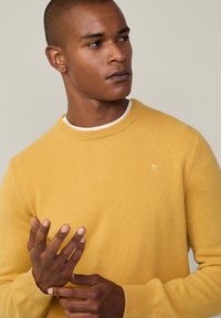 Muž v hořčicově žlutém pleteném svetru s malým emblémem, dívající se stranou a upravující si levé zápěstí pravou rukou.