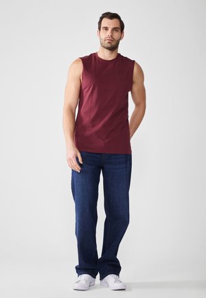 Bordo bezrukavi top od pamuka, uparen s plavim trapericama. Model stoji protiv jednostavne pozadine, noseći bijele tenisice.