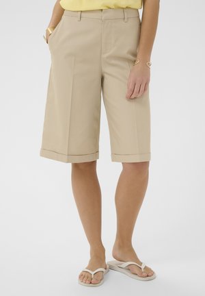 Femme portant un short beige cintré jusqu'aux genoux, un haut jaune, des bracelets dorés et des tongs blanches, debout les mains détendues.