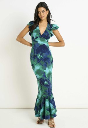 Woman wearing a long, sleeveless green and blue floral dress with ruffled shoulders and hem, posing with hands on hips against a white background.