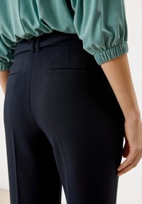 Marineblauwe op maat gemaakte broek gezien vanaf de achterkant, met twee achterzakken en een slanke fit. Het bijpassende bovenstuk is lichtgroenblauw met gathered mouwen.