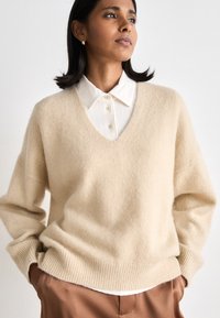 Beige wool v-neck sweater with dropped shoulders and ribbed hem, layered over a white collared shirt with button detailing.