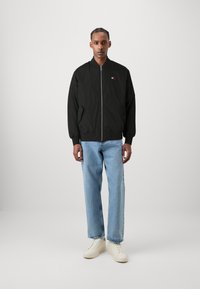 Svart bomberjakke med glidelås foran og sidelommer, brukt over lyseblå rette jeans og kremfargede sneakers. Ensfarget, lett stoff.