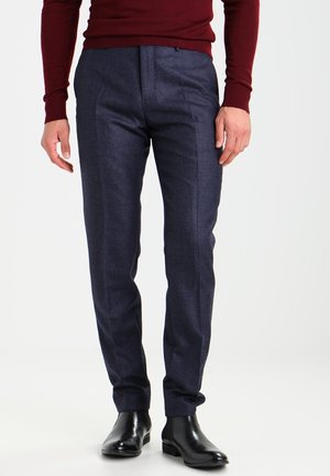 Man draagt donkerblauwe geruite nette broek, zwarte leren nette schoenen en een bordeauxrode aansluitende trui met lange mouwen.