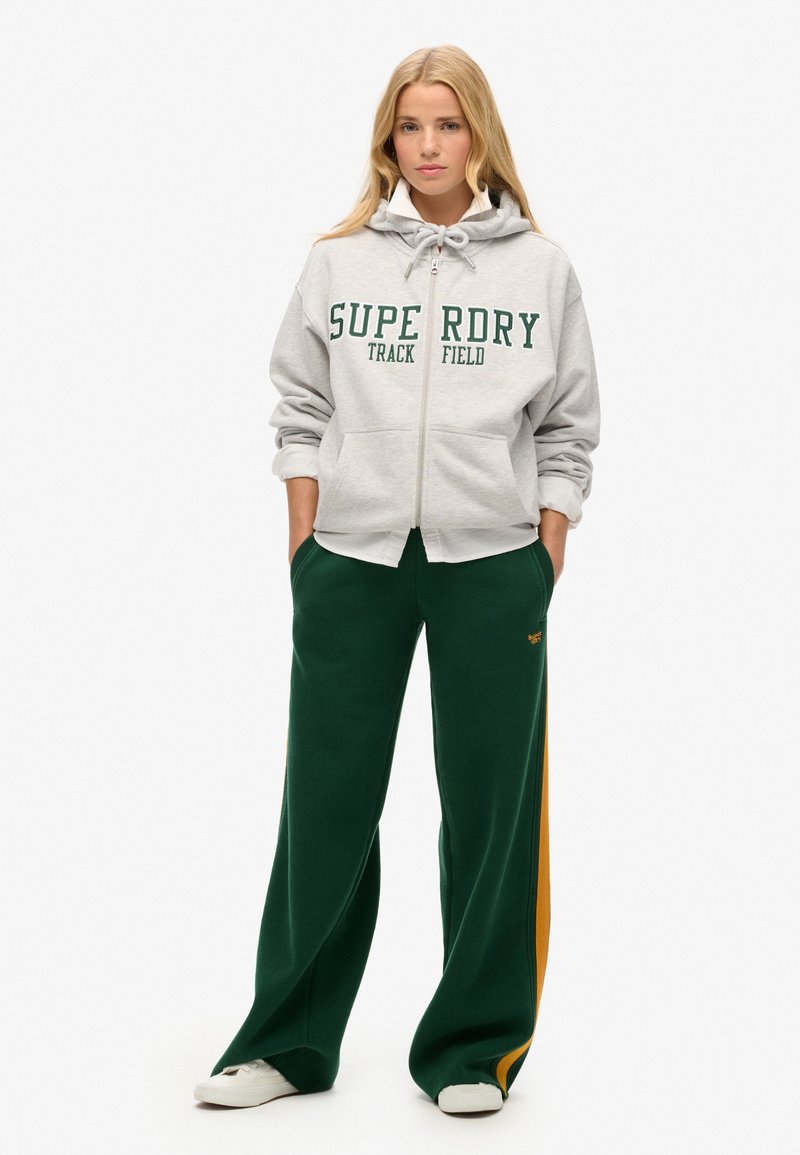 Grå zip-up hoodie med "SUPERDRY TRACK FIELD" tryk i grøn, parret med mørkegrønne sweatpants med et gult sideløb.