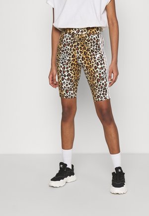 Personne portant un short cycliste imprimé léopard avec des bandes blanches sur les côtés, des chaussettes blanches, des baskets noires et blanches, et un t-shirt blanc.