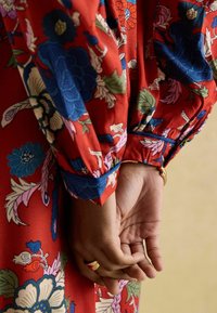 Rotes Blumenkleid mit blauen und rosa Mustern, mit Puffärmeln und gerafften Manschetten. Die Hände sind hinter dem Rücken gefaltet, wobei ein goldener Ring sichtbar ist.