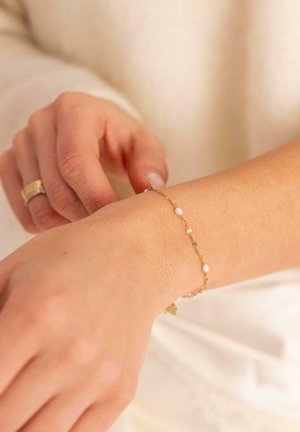 Delicate gold chain bracelet with small pearls worn on left wrist, hand adjusting it, person wearing cream-colored long sleeve top.
