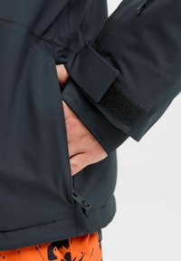 Juodas vandeniui atsparus džemperis, turintis priekinius kišenes, reguliuojamus rankogalius su Velcro ir virvutes juosmens. Lygus paviršius su elegantišku dizainu.