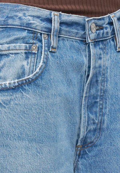 Κοντινή λήψη ανοιχτό μπλε τζιν παντελονιού με μεταλλικό κουμπί και τρουκς, μπροστινή τσέπη και καφέ ριγωτό κορσάζ που έχει μπει μέσα.