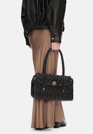 Persona con giacca di pelle nera e gonna marrone che tiene una borsa nera con texture e dettagli in fibbia dorata alle maniglie.