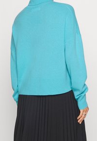 Pull en tricot bleu clair avec un col haut, des épaules tombantes et des poignets côtelés. Associé à une jupe plissée noire.