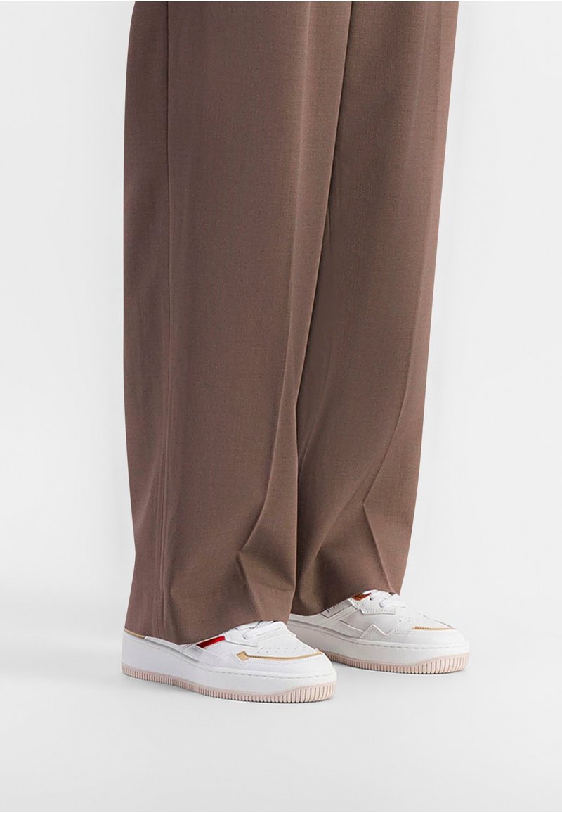 Witte sneakers met textuurdetails en roze rubberen zolen, gecombineerd met lange bruine broeken van gladde stof, die de schoenen gedeeltelijk verbergen.