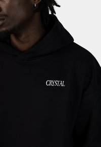 Felpa nera con un tessuto morbido, caratterizzata da un logo "CRYSTAL" ricamato in bianco sul petto. Design con cappuccio e vestibilità rilassata.