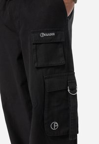 Black cargo pants with multiple side pockets, a metal ring, and embroidered logo. Made of sturdy fabric with a smooth texture.