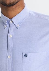 Ljust blå skjorta med knappar tillverkad av strukturerad textil, med en bröstficka som har en broderad logotyp och vita knappar.