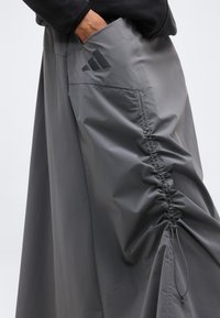 Χέρι στην τσέπη γκρι παντελονιών Adidas με ρυτιδώσεις και μαύρο λογότυπο στο πλάι, φορεμένα με μαύρο μακρυμάνικο τοπ.