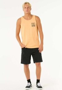 Oranges Baumwoll-Tanktop mit schwarzem Logo auf der Brust, kombiniert mit schwarzen Denim-Shorts und weißen Sneakers mit schwarzen Akzenten.