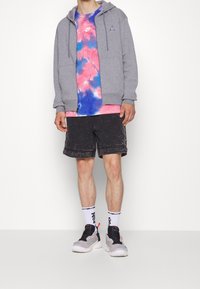 Felpa grigia con zip, T-shirt multicolore tie-dye, pantaloni corti neri con risvolti e sneakers grigie con dettagli. Calzini bianchi con design nero.