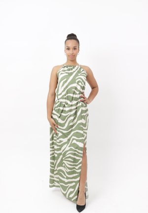 Vrouw met haar in een knot, die een lange groene en witte jurk met zebraprint en een zijsplit draagt en zwarte hakken, staand met één hand op haar heup.