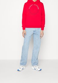 Red hoodie with “Tommy Hilfiger New York City” print, paired with light blue jeans and white sneakers with blue accents.