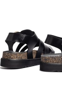 Sandalias de cuero negro con tiras cruzadas, plantilla de corcho, plataforma texturizada y un ligero tacón. Presentan un acabado suave y un diseño duradero.