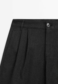 Gros plan sur un pantalon plissé gris foncé avec passants de ceinture, poche latérale et fermeture à bouton noir à la taille.