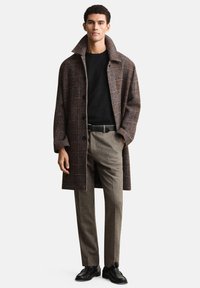 Manteau à carreaux marron avec un col montant, superposé sur un pull noir, associé à un pantalon gris et des chaussures en cuir noires.