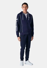 Felpa con zip color navy, con logo bianco e dettagli in piping, abbinata a pantaloni jogger coordinati. Il materiale sembra morbido. Il modello indossa sneakers bianche.