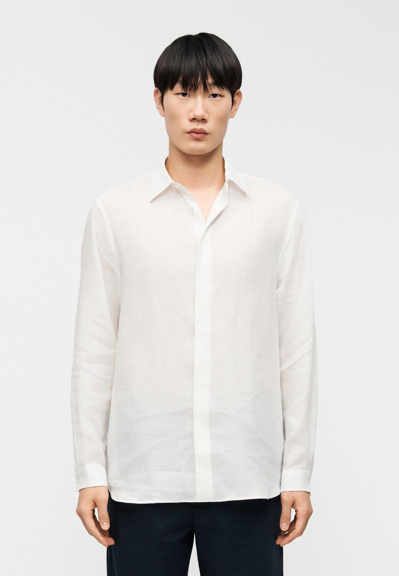 Chemise blanche à manches longues en tissu léger, avec col, boutons à l'avant et coupe droite. Texture lisse avec peu de plis.