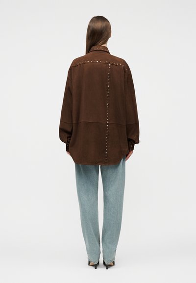 Γυναίκα που φοράει χαλαρό καφέ σακάκι με τρουκς και ανοιχτό μπλε τζιν, στέκεται με την πλάτη στην κάμερα σε απλό λευκό φόντο.