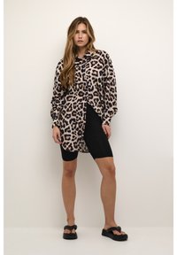Camisa de botón de gran tamaño con estampado de leopardo en beige claro y marrón oscuro, con mangas largas y umbral asimétrico. Usada sobre pantalones cortos de ciclismo negros.