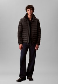 Must pufferjakk horisontaalse tikandiga, luku ja kõrge kraega, kantud musta lukuga topi ja tumesiniste teksade peal.