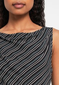 Közelkép egy olyan személyről, aki ujjatlan fekete felsőt visel fehér és szürke függőleges hullámos csíkokkal, látható a nyaka, ajkai és a mellkasa felső része.