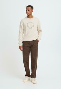 Beige sweatshirt med cirkulært broderet tekst, parret med mørkebrune bukser og cremefarvede sneakers. Modellen står foran en lys baggrund.