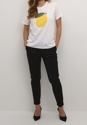 Donna che indossa una maglietta bianca con una grafica di limone giallo, pantaloni neri slim e tacchi neri con punta, in piedi con le mani in tasca.