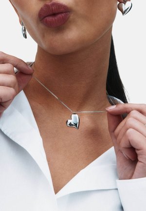 Collana con ciondolo a forma di cuore in argento, con superficie liscia e riflettente, attaccata a una catena delicata. Modello che indossa una camicia bianca.