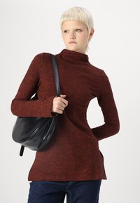 Pulover închis cu guler înalt, cu mâneci lungi, dintr-un material tricotat texturat. Geanta neagră cu formă rotunjită, ținută în mâna stângă, purtată cu blugi albaștri.