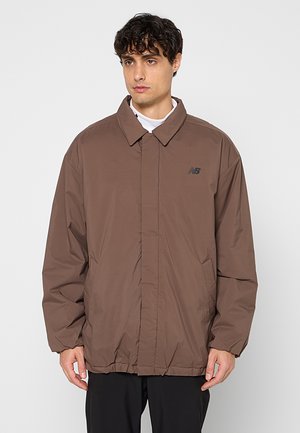 Mann trägt eine braune New Balance gesteppte Jacke mit Kragen, Fronttaschen und verdecktem Reißverschluss, steht vor einfachem Hintergrund.