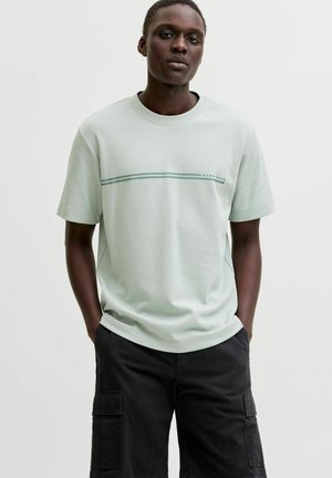 Fiatal, sötét bőrű férfi világoszöld, rövid ujjú ingben, fekete cargo rövidnadrággal, kezét a zsebekben tartva.