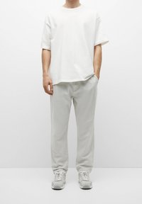 T-shirt bianca in cotone con maniche corte, abbinata a pantaloni grigi chiari e larghi e sneakers grigie, in piedi contro uno sfondo neutro.