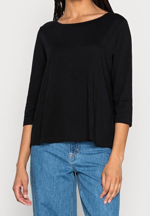 Femme portant un haut noir ample à manches trois-quarts et un jean bleu taille haute, sur un fond uni.