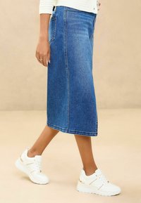 Denim midi sukně s rovný střih, světle modré barvy, zadními kapsami a bílými teniskami se zlatými akcenty. Texturovaná látka.
