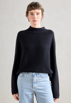 Pull en maille noir à col montant, avec des manches larges et une coupe décontractée, associé à un jean en denim bleu clair. Texture douce, design minimaliste.